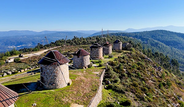 Imagem de lindos moinhos de vento históricos em uma colina com vista para a paisagem natural, céu azul e uma vista panorâmica de montanhas ao fundo, destacando a beleza e a preservação da arquitetura tradicional em um cenário rural.