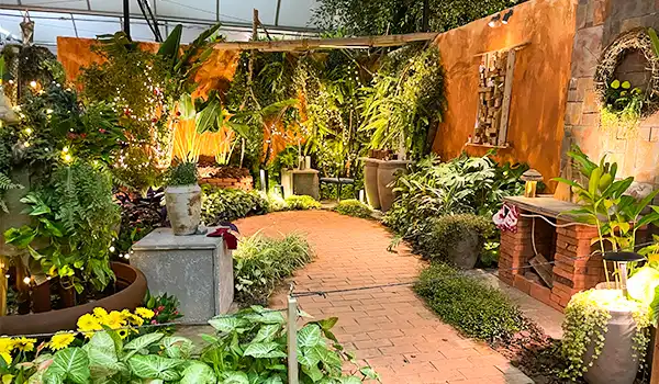 Jardim com plantas variadas, caminho de tijolos, paredes decoradas e iluminação suave criando um ambiente tranquilo e natural ao ar livre. Parada de Natal de Holambra.