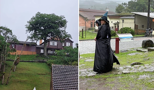 Imagem de uma pessoa usando capa de chuva preta e chapéu em um ambiente urbano com pedras e árvores ao fundo, sob chuva, em um dia chuvoso.