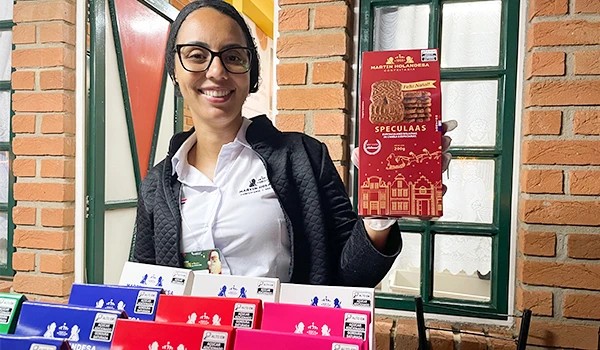 Mulher sorridente segurando uma caixa de chocolates "Speculas" em uma loja, fundo de parede de tijolos e janela verde. Produto de alta qualidade em destaque.