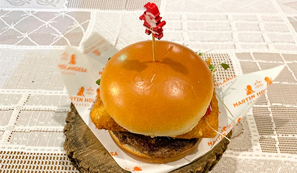 Sanduíche gourmet com hambúrguer suculento, pão fofinho e uma fatia de burger de queijo, decorado com um palito vermelho e servido sobre fatia de tronco de madeira.