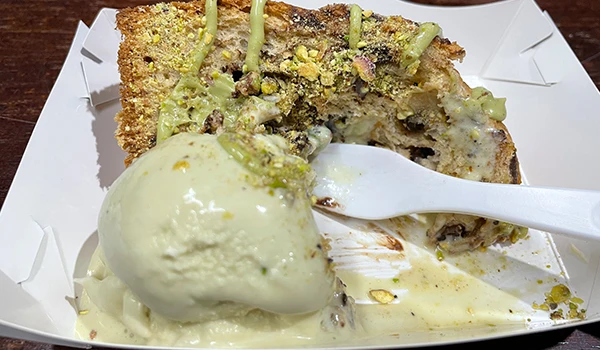 Sobremesa de pudim de pistache com calda de chantilly e castanhas, apresentada em uma bandeja de papel.