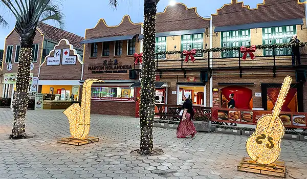 Imagem de uma praça decorada com luzes de Natal, incluindo árvores iluminadas e relíquias de instrumentos musicais em tamanho gigante, com uma loja de doces ao fundo. Parada de Natal de Holambra.