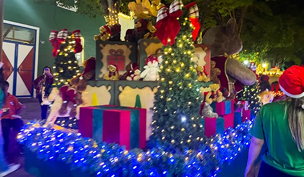 Decoração de Natal com árvore decorada, ursinhos de pelúcia, presentes coloridos, luzes natalinas e figuras festivas em um cenário exterior, criando uma atmosfera natalina encantadora.