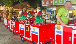 Família participando de um passeio natalino com carrinho de Natal decorado, coxinhas e figurinos de Natal, em ambiente externo durante a noite.