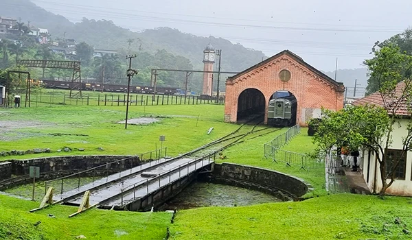 Estação de trem histórica rodeada por uma paisagem verdejante e montanhas ao fundo, com um caminho de trilho de trem levando até um prédio antigo de tijolos.