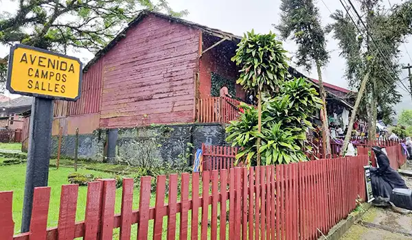 Casa simples localizada na avenida Campos Salles, com acabamento de madeira, cercada por um muro vermelho e rodeada de árvores e vegetação.