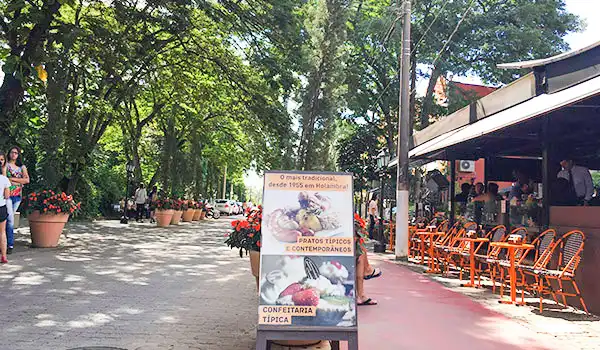 Ambiente de rua em uma rua de lazer com árvores, mesas ao ar livre e um cardápio de confeitaria tipica, incluindo aves e flores para decoração.