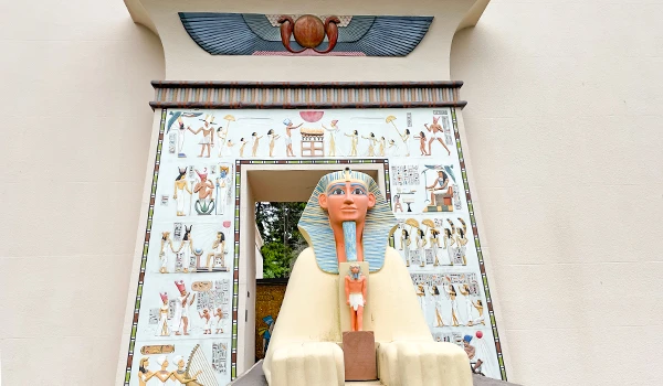 Imagem de uma atração temática com um grande busto na entrada, decorada com arte egípcia e hieróglifos, em um parque ou museu com decoração inspirada no Egito antigo.
