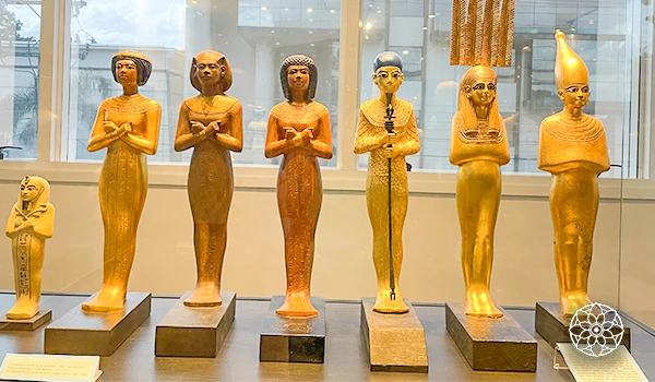 Conjunto de estátuas de deuses egípcios antigos, todas de cor dourada, exibindo diferentes poses e atributos tradicionais, expostas em um museu com fundo de janela.