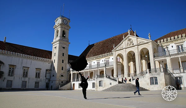 Praça histórica com prédio antigo e torre, céu claro ao fundo, pessoas caminhando na frente, arquitetura colonial.