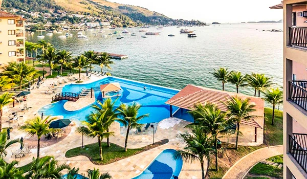 Vista de uma piscina com formato de peixe em um resort à beira-mar, rodeada por palmeiras e com vista para o oceano, apresentando um cenário de descanso e lazer. Hotéis da Costa Verde do Rio de Janeiro.