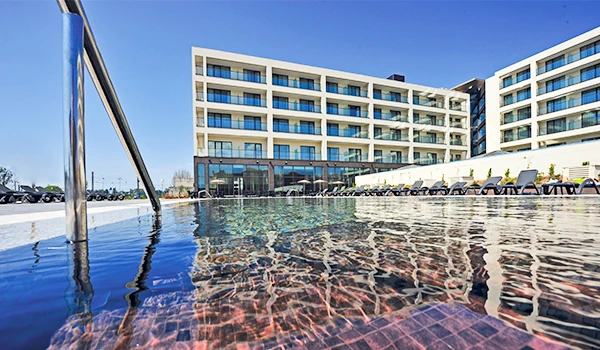 Vista de um hotel com piscina de borda infinita, com estrutura moderna de vários andares e espreguiçadeiras ao redor, sob céu azul claro.