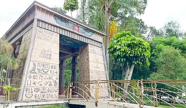 Imagem de um monumento inspirado na arquitetura egípcia antiga, com hieróglifos nas paredes, localizado em meio a uma área verde com árvores. Perfeito para quem busca referências de construções temáticas ou culturais.