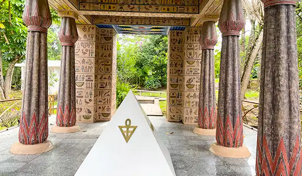 Estrutura com pilares decorativos e hieróglifos, em um ambiente ao ar livre, com uma pirâmide de granito ao centro, rodeada de natureza verde.