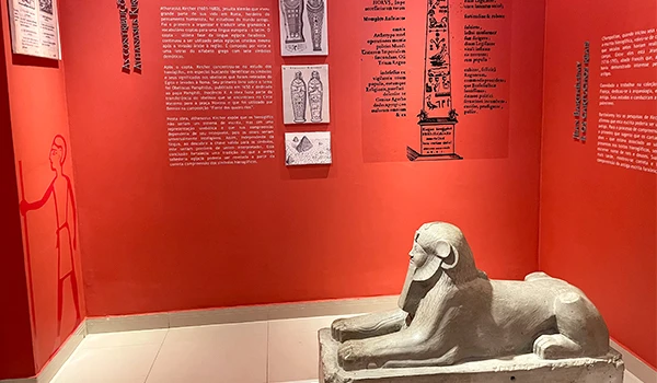 Estátua de um leão egípcio antiga exibida em um museu com paredes vermelhas e textos históricos ao fundo. Museu Egípcio e Rosacruz em Curitiba.