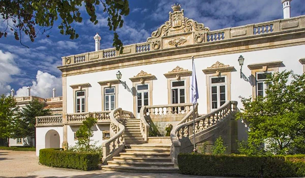 Imagem de uma bela mansão histórica com escadaria imponente, fachada branca, detalhes ornamentais no topo e ao redor, cercada por árvores verdes e céu azul com nuvens.