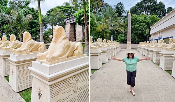 Imagem de uma Deusa Rodrigues visitando uma exposição de réplicas de esculturas de antigos faraós egípcios ao ar livre, com muitos bustos alinhados ao longo do caminho e um cenário de natureza ao fundo.