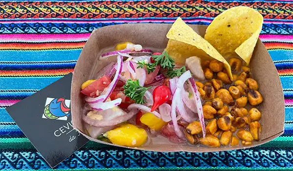 Salada de cebola, milho, tomate, e nachos em uma embalagem de papel, tradicional na culinária mexicana, disposta sobre uma mesa com tapete colorido.