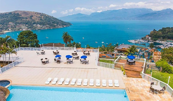 Vista panorâmica de uma piscina de hotel com cadeiras de sombra azuis, cercada por uma varanda com vista para um lago e montanhas ao fundo, em um dia ensolarado. Hotéis da Costa Verde do Rio de Janeiro.