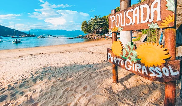 Praia de Porto Girasol com sinalização de pousada ao lado da areia, vista para o mar e barcos ao fundo em uma manhã ensolarada.