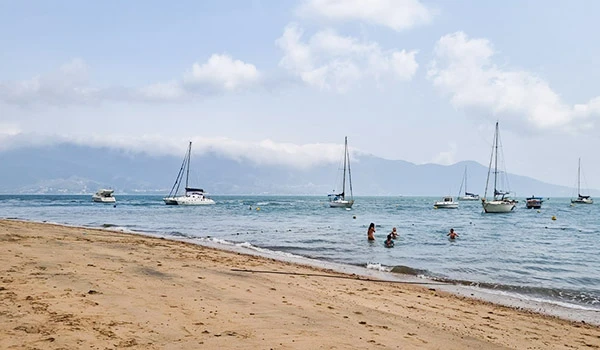 Praia com barcos à vela ao fundo, areia dourada, pessoas nadando e céu parcialmente nublado, cenário perfeito para um dia de descanso. Ilhabela turismo e casamentos.