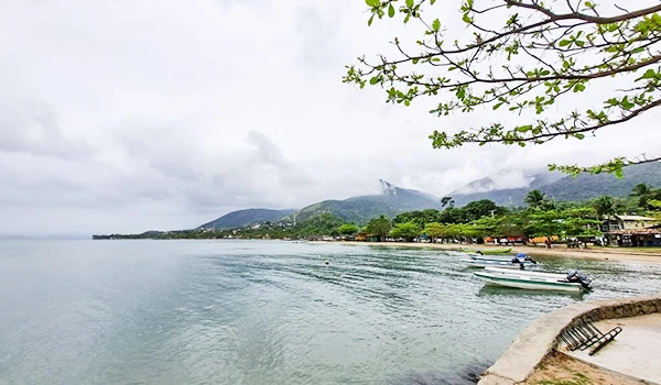 Praia tranquila em uma cidade litorânea com um barco ancorado, árvores verdes e montanhas ao fundo em um dia nublado. Ilhabela turismo e casamentos.