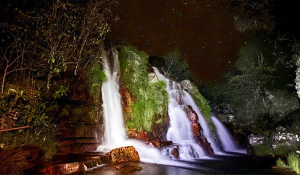 Cachoeira iluminada à noite com águas caindo por rochas e vegetação ao redor, criando um cenário natural e relaxante em uma floresta. Turismo na Chapada dos Veadeiros.