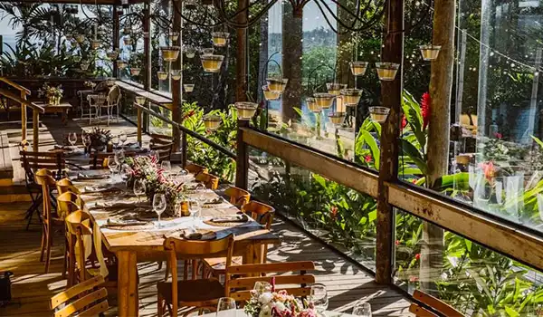 Ambiente rústico de restaurante com decoração charmosa, mesas bem postas com guardanapos de pano, flores e cadeiras de madeira, rodeado por plantas e iluminação natural.