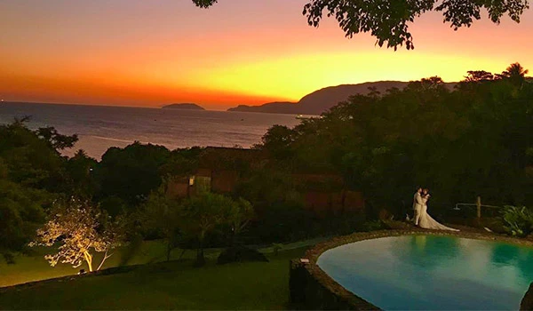 Pôr do sol deslumbrante sobre o oceano, com céu em tons de laranja e rosa, rodeado por árvores e uma piscina ao lado de um casal, criando cenário de beleza natural e tranquilidade.