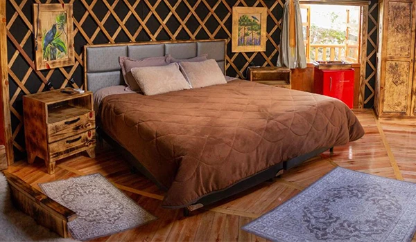 Quarto aconchegante com cama de casal, cabeceira estofada, cortinas, tapetes e decoração rústica com detalhes de madeira e pintura artística, ideal para relaxar.