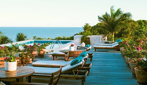 Terraço de um hotel com vista para o mar, lounge ao ar livre com espreguiçadeiras, mesas de madeira e muitas plantas ornamentais em um clima ensolarado, ideal para relaxar e aproveitar a natureza.
