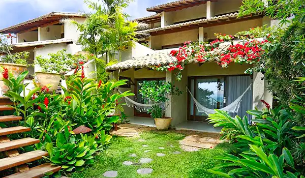 Imagem de uma pousada charmosa com fachada de cores claras, varanda com rede, jardim bem cuidado com plantas e flores, ideal para férias relaxantes na natureza.