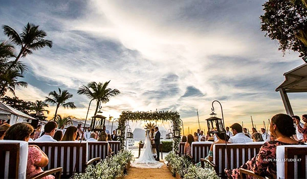 Cerimônia de casamento ao pôr do sol ao ar livre com convidados, decoração romântica com flores e iluminação aconchegante na praia. Ilhabela turismo e casamentos.