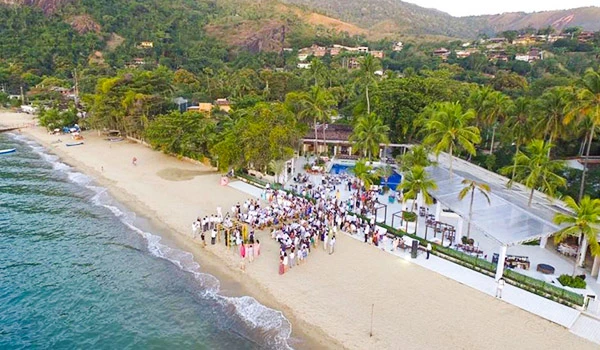 Vista aérea de uma cerimônia de casamento na praia de Ilhabela com muitas pessoas, cercada por palmeiras e áreas verdes na orla de uma cidade costeira.