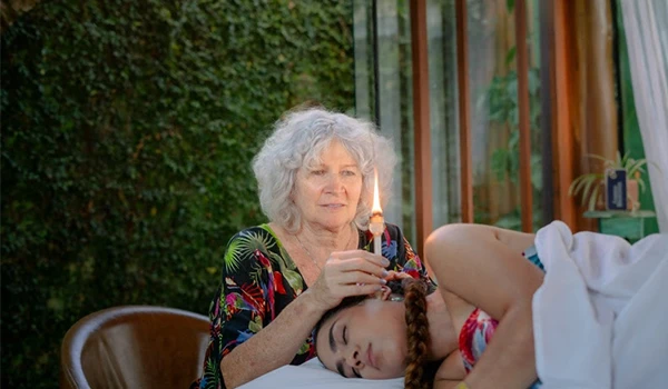 Cena de uma mulher idosa cuidando de uma jovem deitada, em ambiente interno com janelas de vidro e vegetação ao fundo, representando cuidado e afeto.