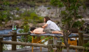 Sessão de massagem ao ar livre com uma mulher recebendo massagem relaxante enquanto deita em uma cama portátil em meio à natureza ao redor de árvores e rio.
