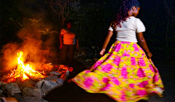Mulher dançando ao redor de uma fogueira na noite, vestida com saia colorida e camisa branca, em um ambiente ao ar livre com árvores ao fundo. Turismo na Chapada dos Veadeiros.