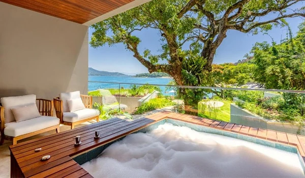 Terraço com banheira de hidromassagem ao ar livre, vista para a natureza, árvores e o mar ao fundo, ambiente de relaxamento e lazer no exterior.