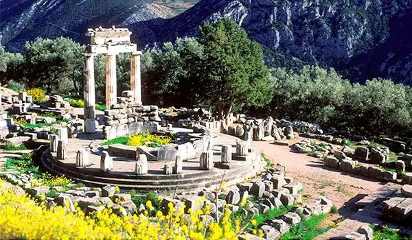 Ruínas antiguas na encosta de uma colina com colunas de pedra e restos de construções, cercadas por vegetação e árvores, sob céu azul claro.