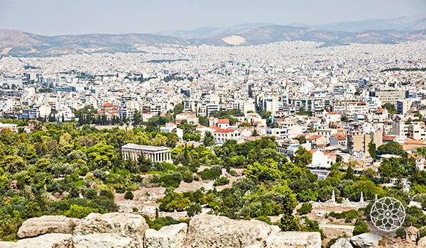 Imagem panorâmica de Atenas, mostrando o Monte Licabeto cercado por árvores verdes e o horizonte urbano com edifícios variados ao fundo, sob céu claro.