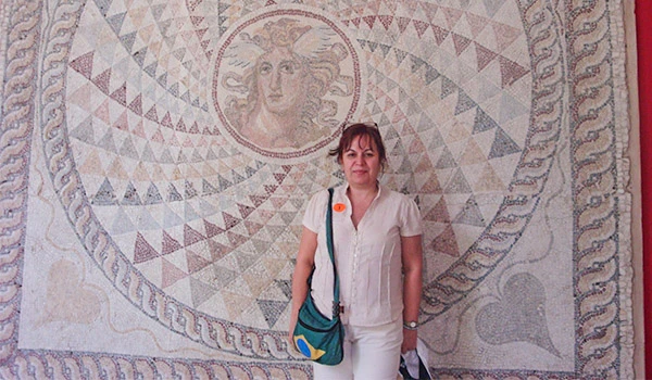 Deusa Rodrigues posando diante de uma antiga mosaico de Teseu com Minotauro, com detalhes geométricos em cores suaves no fundo. Onde ficar na Grécia.