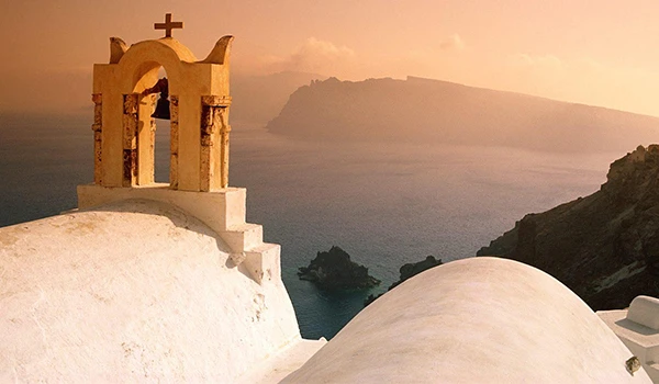 Cenário costeiro de Santorini ao pôr do sol, com uma capela branca tradicional grega em primeiro plano, destacando o pôr do sol e o mar azul ao fundo.