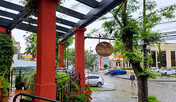 Imagem de uma rua na cidade de Madalena, com árvores, carros estacionados e uma loja chamada "Velho Madaloso" ao fundo, em um dia nublado. Perfeito para quem busca dicas de turismo e gastronomia na região de Madalena, com ambiente acolhedor e belas paisagens urbanas.