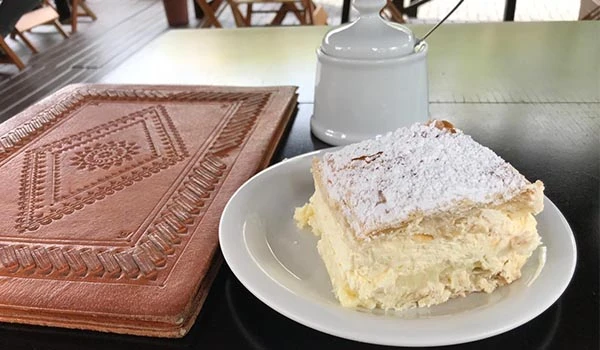 Fatia de bolo com creme e cobertura de farinha de trigo, servida em prato branco, ao lado de uma xícara e uma panela de leite, em uma mesa de madeira.