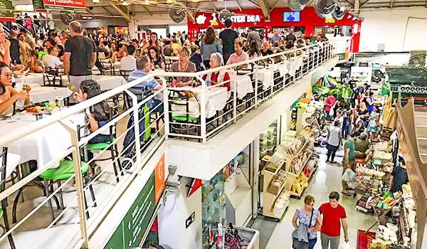 Imagem de um shopping movimentado com várias lojas, pessoas fazendo compras e comendo em um restaurante no andar superior, mostrando um ambiente comercial vibrante.