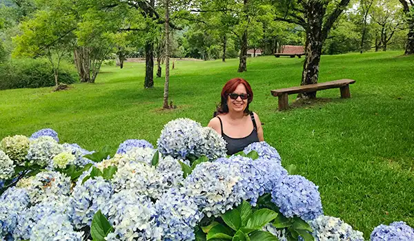 Deusa Rodrigues ao lado de flores de hortênsia azul e branca em parque verde com árvores ao fundo, dia ensolarado.