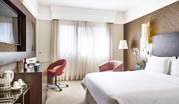 Quarto de hotel confortável e bem decorado com cama de casal, televisão, escrivaninha, cadeira de escritório, poltrona, luminária e cortinas de tecido claro.