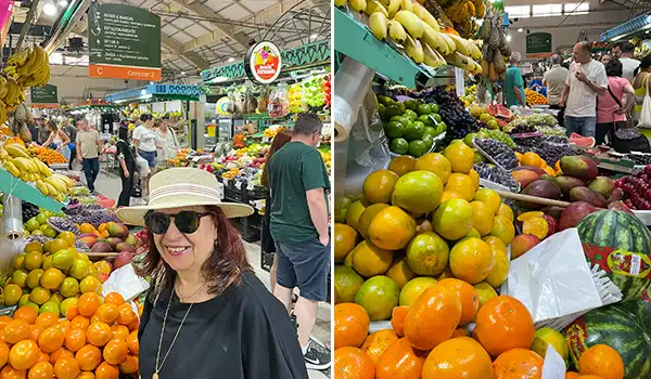 Mulher com chapéu, óculos escuros e blusa preta em feira livre com diversas frutas ao fundo, incluindo bananas, laranjas, melancias e uvas, durante compras de frutas frescas.