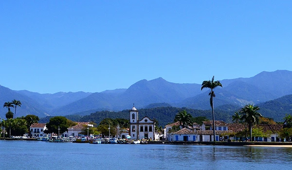 Paisagem de uma cidade costeira com lago, igrejas tradicionais, palmeiras e montanhas ao fundo, em um dia ensolarado no Brasil.